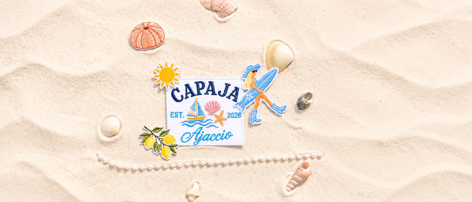 Contact Patch brodé Capaja, Ajaccio, Est. 2026, posé sur le sable avec d'autres patchs, des coquillages et des perles